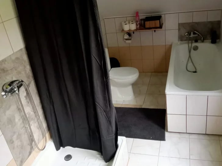 Bathroom interior with black shower curtain, white tiled floor, toilet, bathtub, and wall-mounted fixtures
