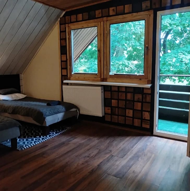 Bedroom with wooden flooring, single bed, and large windows overlooking green foliage
