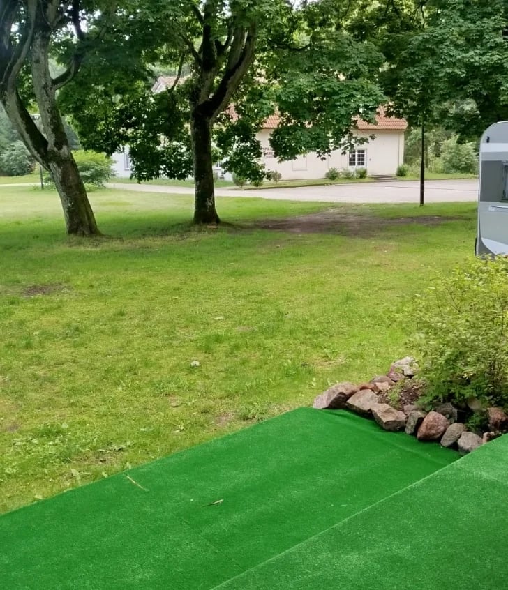 Bright green mini golf course mat with stone border in a park setting with large trees and a building visible in background