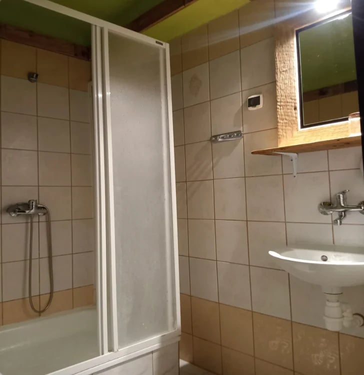 Modern bathroom with beige tiled walls, glass shower enclosure, white sink, and wooden shelf under bright ceiling lighting
