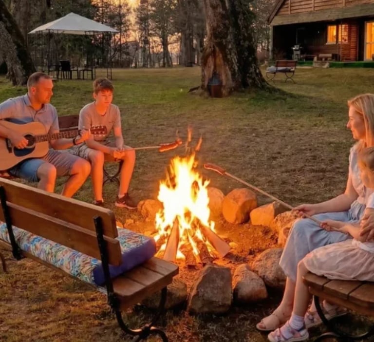 People gathered around a campfire in the evening, with one person playing guitar, sitting on benches in a grassy outdoor area near a cabin