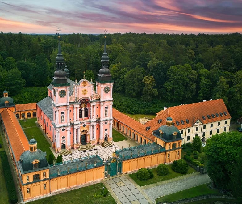 Aerial view of an 18th-century Baroque monastery with pink church and twin spires surrounded by yellow buildings and forest at dusk