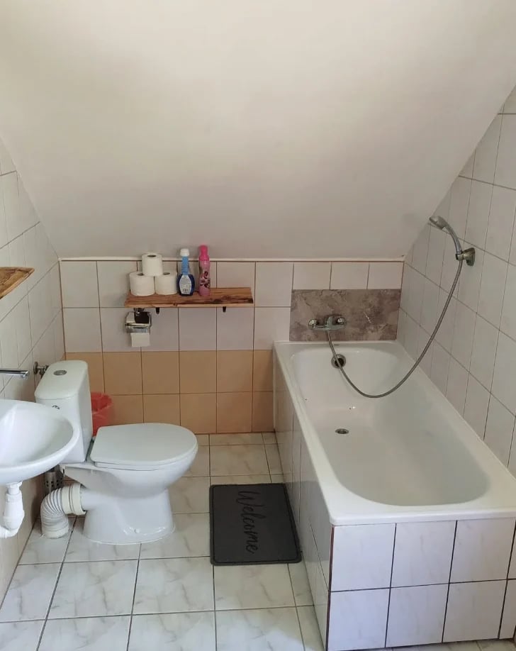 Small bathroom with white toilet, white bathtub, wooden shelf with toiletries, beige and white tile walls, and bathroom mat