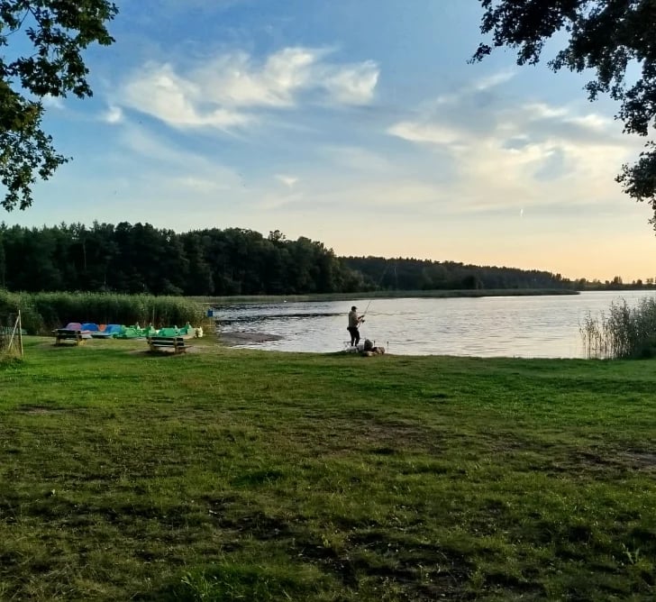 Person fishing on grassy lakeside bank with blue boats, trees and sky reflected in calm water