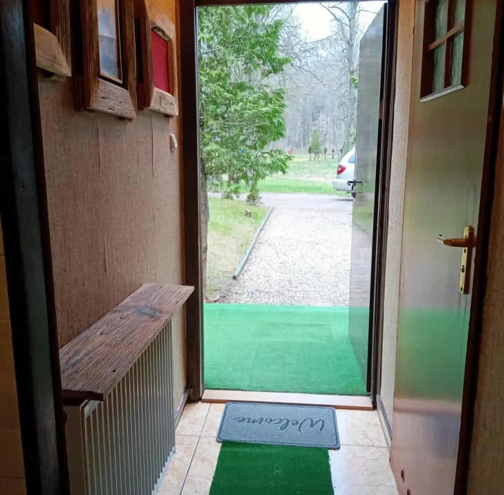 View through an open doorway of a house entrance with green mat, leading to a garden path and trees