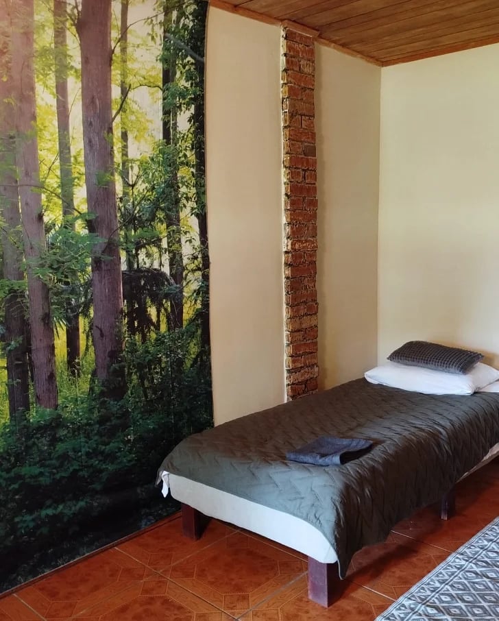 Bedroom with dark bed against exposed brick wall, large forest mural on left wall, wooden ceiling, and terracotta tile floor