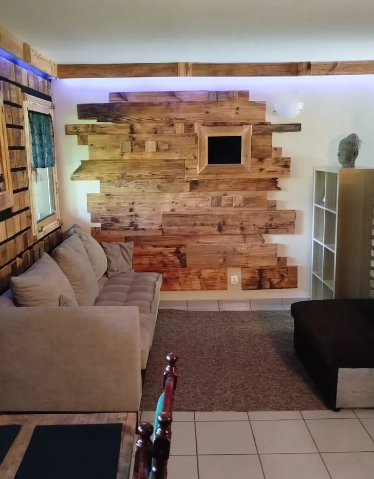Modern living room with a rustic wooden accent wall featuring varied wood planks and a small screen, facing a beige sectional sofa