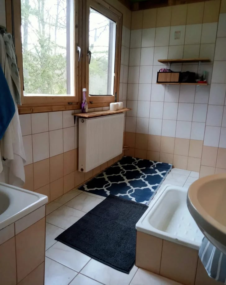 Small bathroom with beige and white tiles, white bathtub, radiator under window, dark blue and black mats on floor, and wall shelving