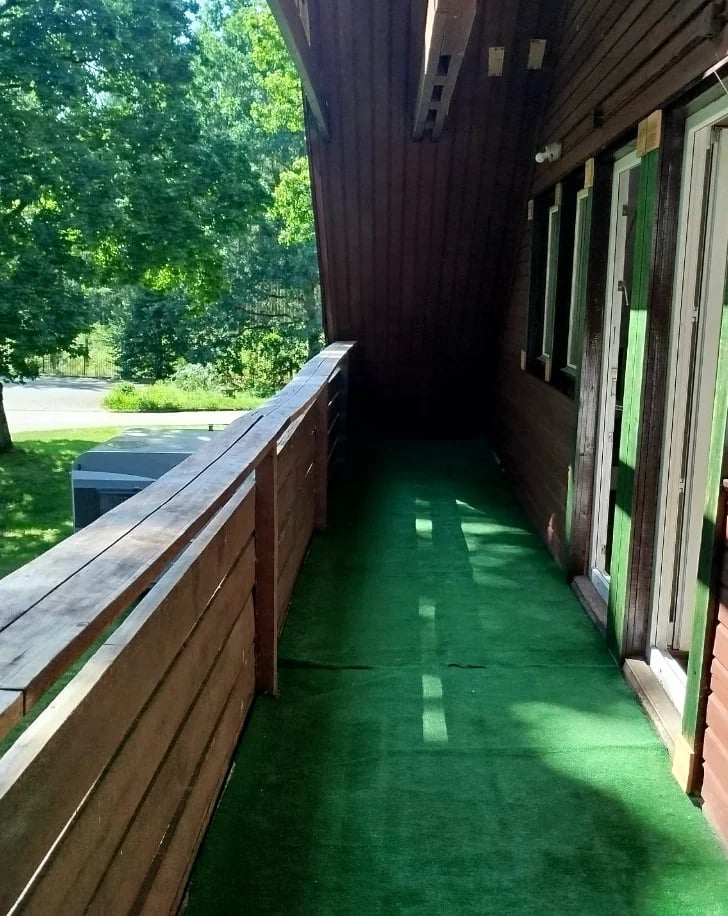 Narrow wooden deck with green painted floor and railings, lined with closed glass doors on the right side, overlooking green trees