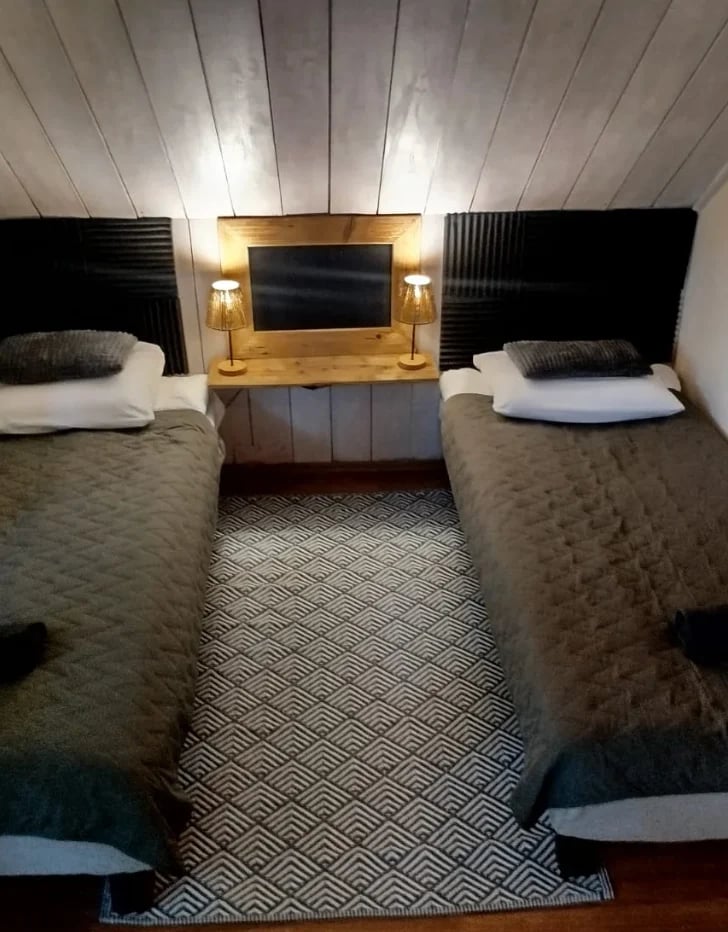 Modern hotel room with two twin beds, wooden accent wall with mounted TV and lamps, geometric floor rug, angled ceiling