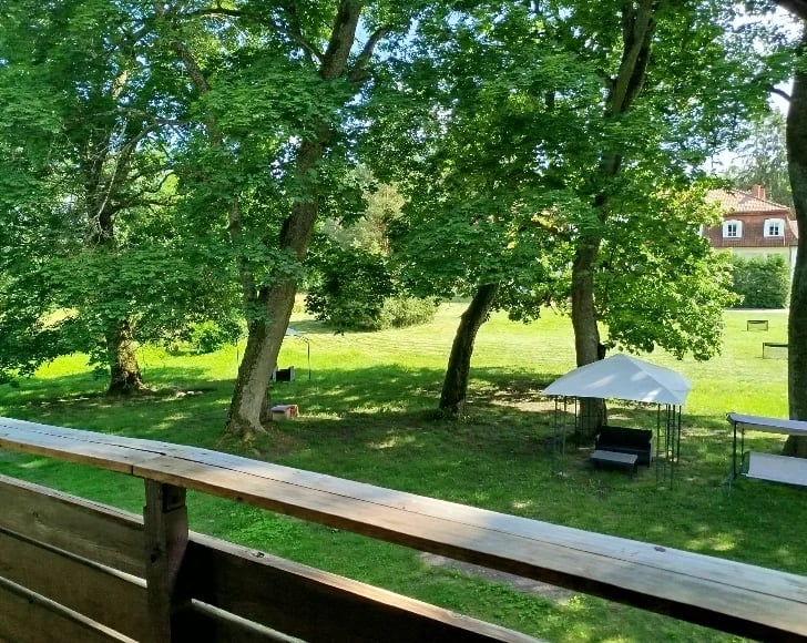Wooden deck railing overlooking a green lawn with large shade trees, picnic table, and house in the background