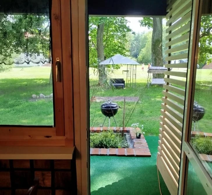 View through wooden doors and louvered shutters of backyard with miniature golf course, grills, and lush green lawn