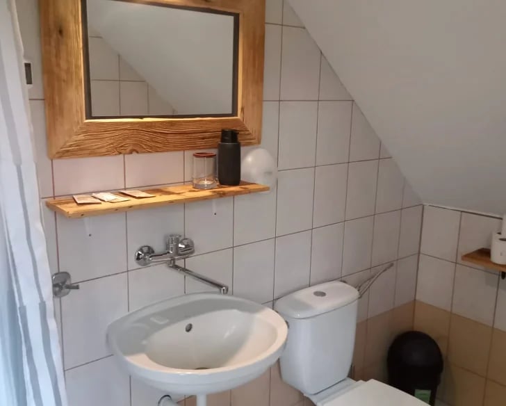 Bathroom with white tile walls, round white sink, wooden mirror frame, shelf with faucet and toilet visible