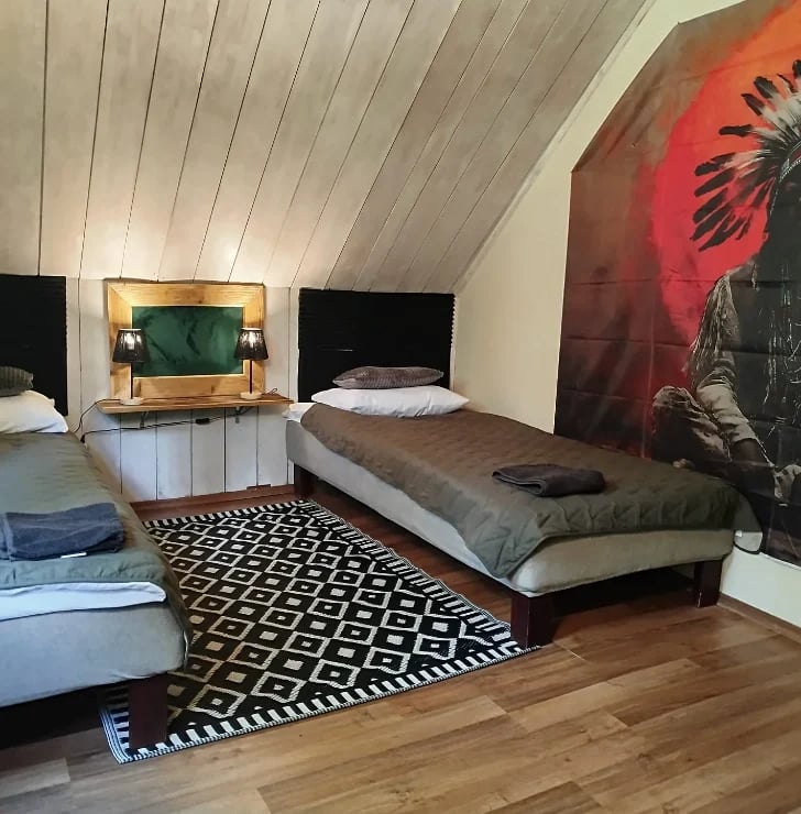 Modern attic bedroom with sloped ceiling, multiple beds, geometric patterned rug, and red wall art featuring a Native American portrait