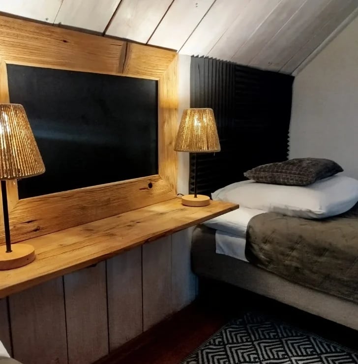 Modern bedroom interior with wooden desk, monitor, and decorative lamps beside a bed with pillows and geometric patterned rug