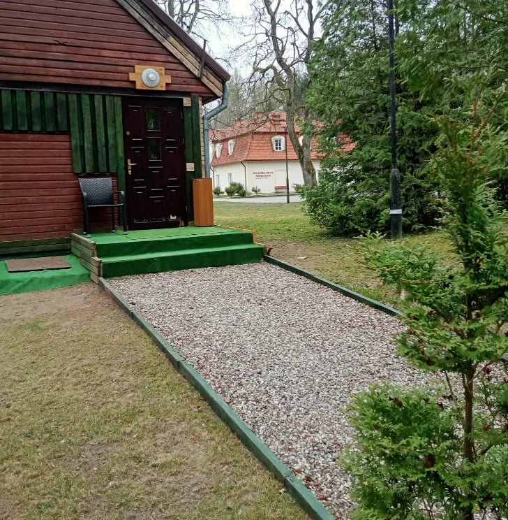 Red wooden cabin with green door and platform, gravel pathway, and green hedges in a garden setting