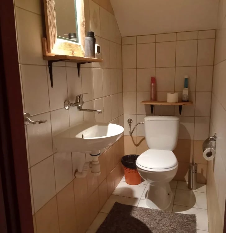 Small bathroom with beige tile walls, white toilet, round sink with mirror above, wooden shelf, and toilet paper holder