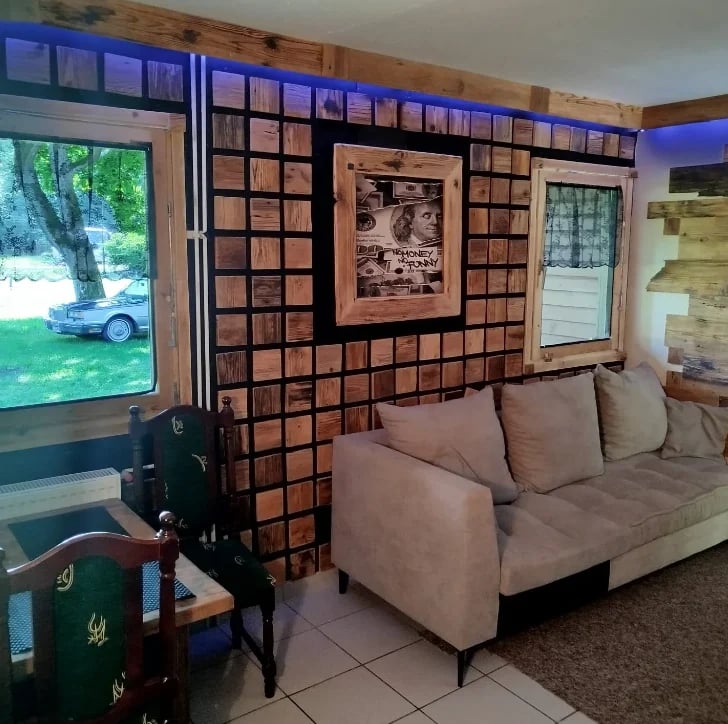 Modern living room with tan sectional sofa, decorative brick wall with framed artwork, large window showing outdoor scenery, and blue-lit ceiling